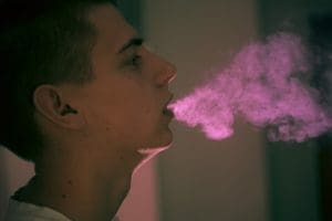 A side profile of a person exhaling a thick cloud of pink vapor in a dimly lit room.